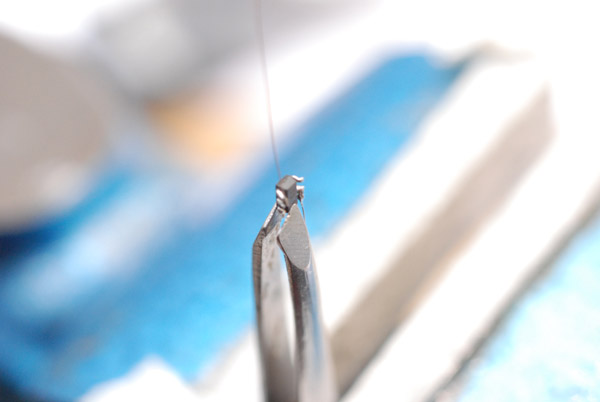 A tiny microchip, held in tweezers, in a vice. Very thin wires soldered to two of the legs