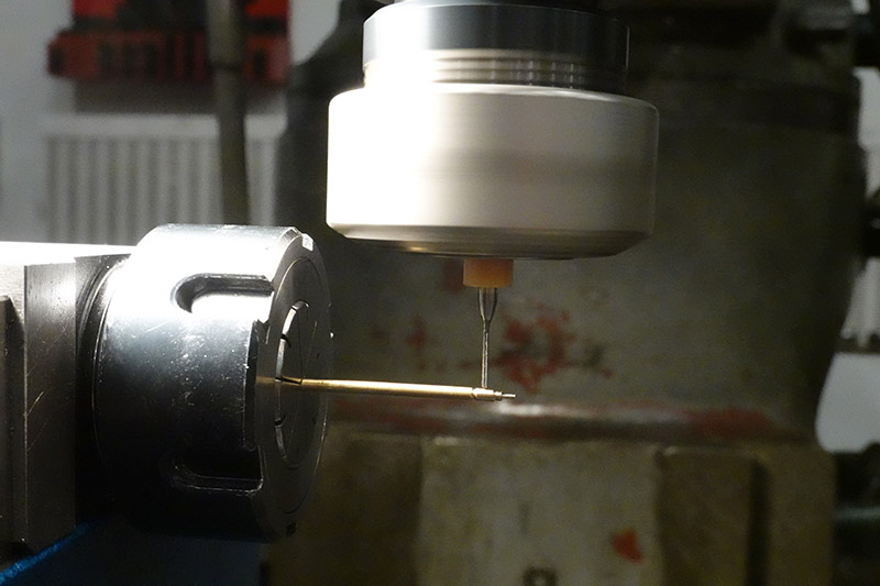 A tiny drillbit held in a collet on the milling machine, approaching the brass stock held in another collet. To stop the collets colliding, the brass stock is extending very far out of the collet.