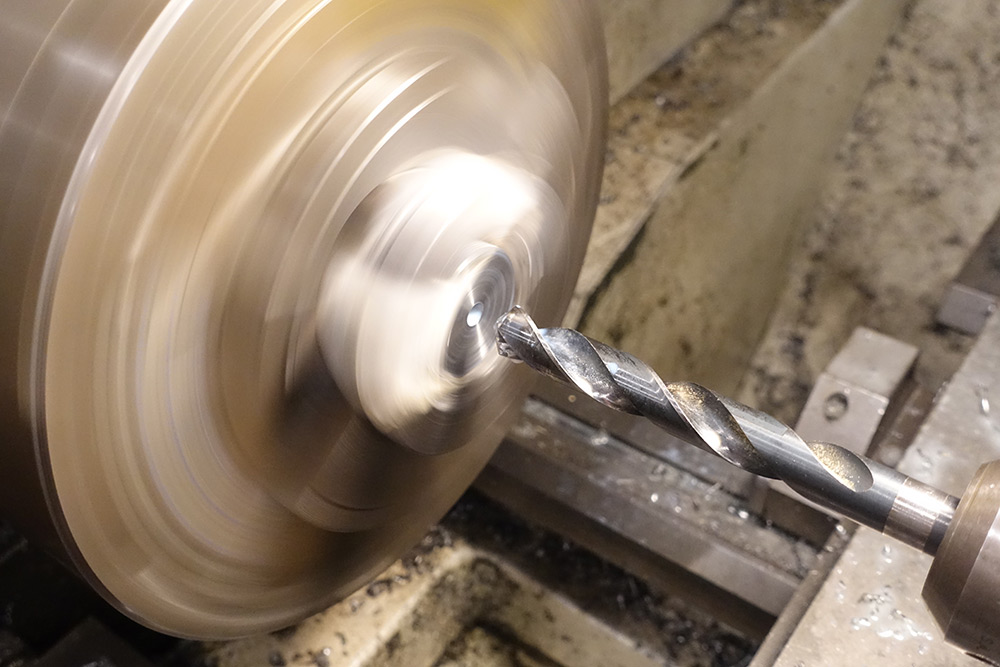 A large drillbit approaches a steel part in a spinning chuck